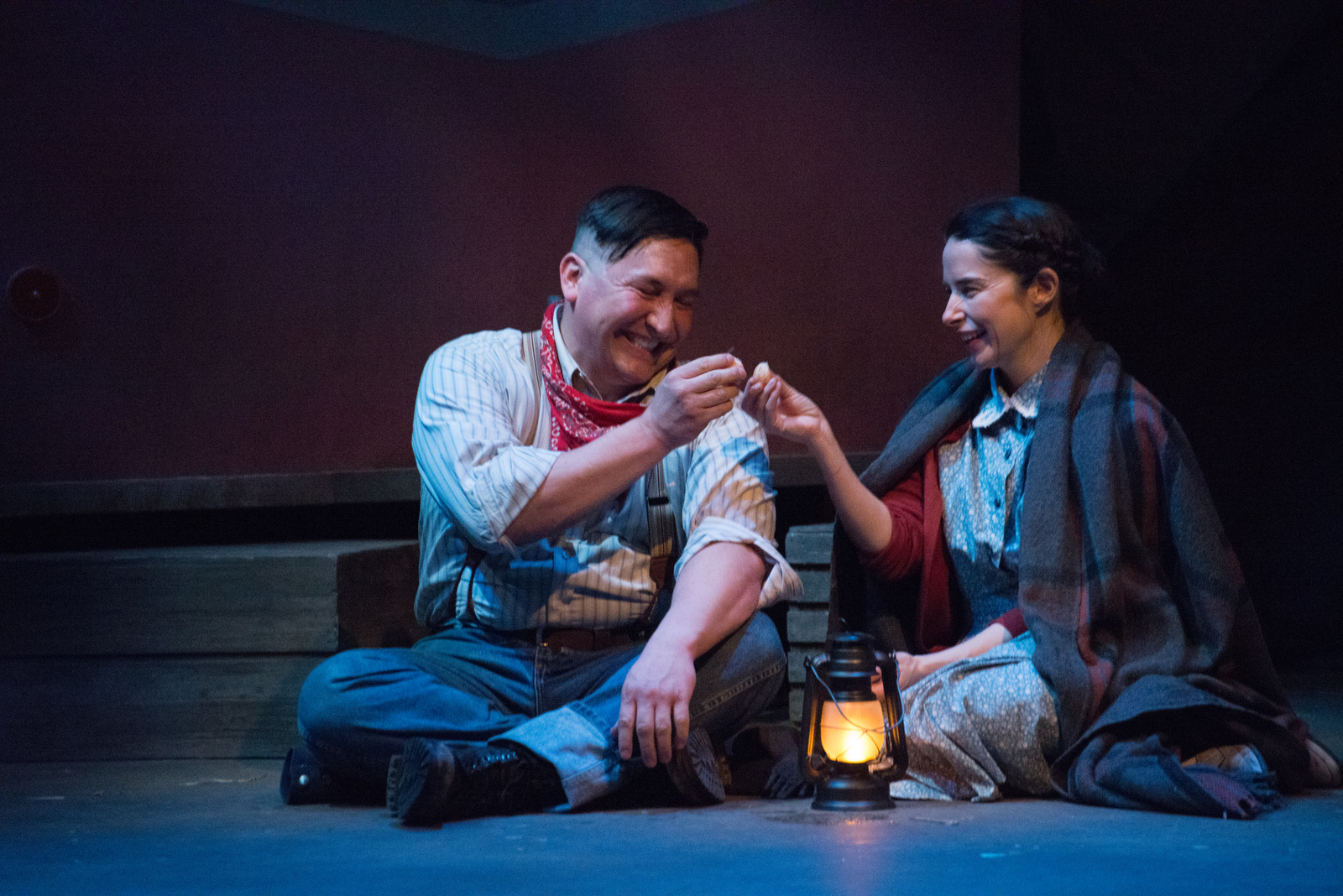In the image, a man and a woman are seated on the ground, engaged in a conversation. The man, dressed in a white shirt and a red bandana, is holding a small object in his hand. The woman, wearing a blue dress, is smiling at him. A lantern is lit in front of them, casting a warm glow on their faces. The background is dark, with a red wall and a wooden door visible.