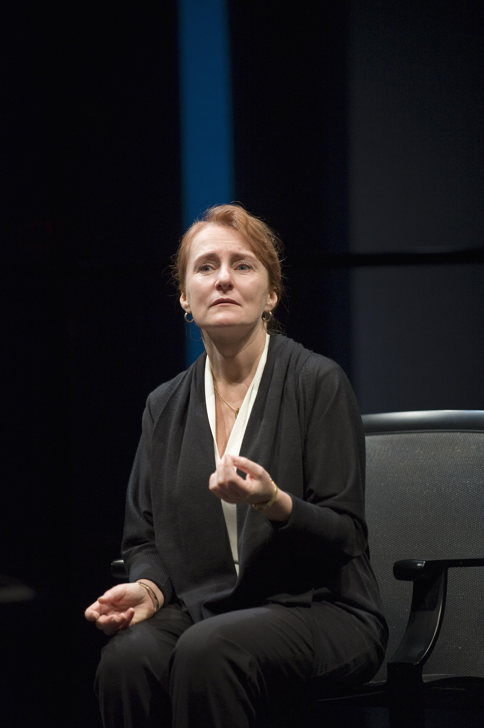 In the image, Actor Seana McKenna is seated in a black chair, her hands gently resting on her lap. She is dressed in a black blazer and white blouse, and her gaze is directed off to the side. The background is dark, providing a stark contrast to the woman's attire. A blue curtain hangs in the background, adding a touch of color to the scene.